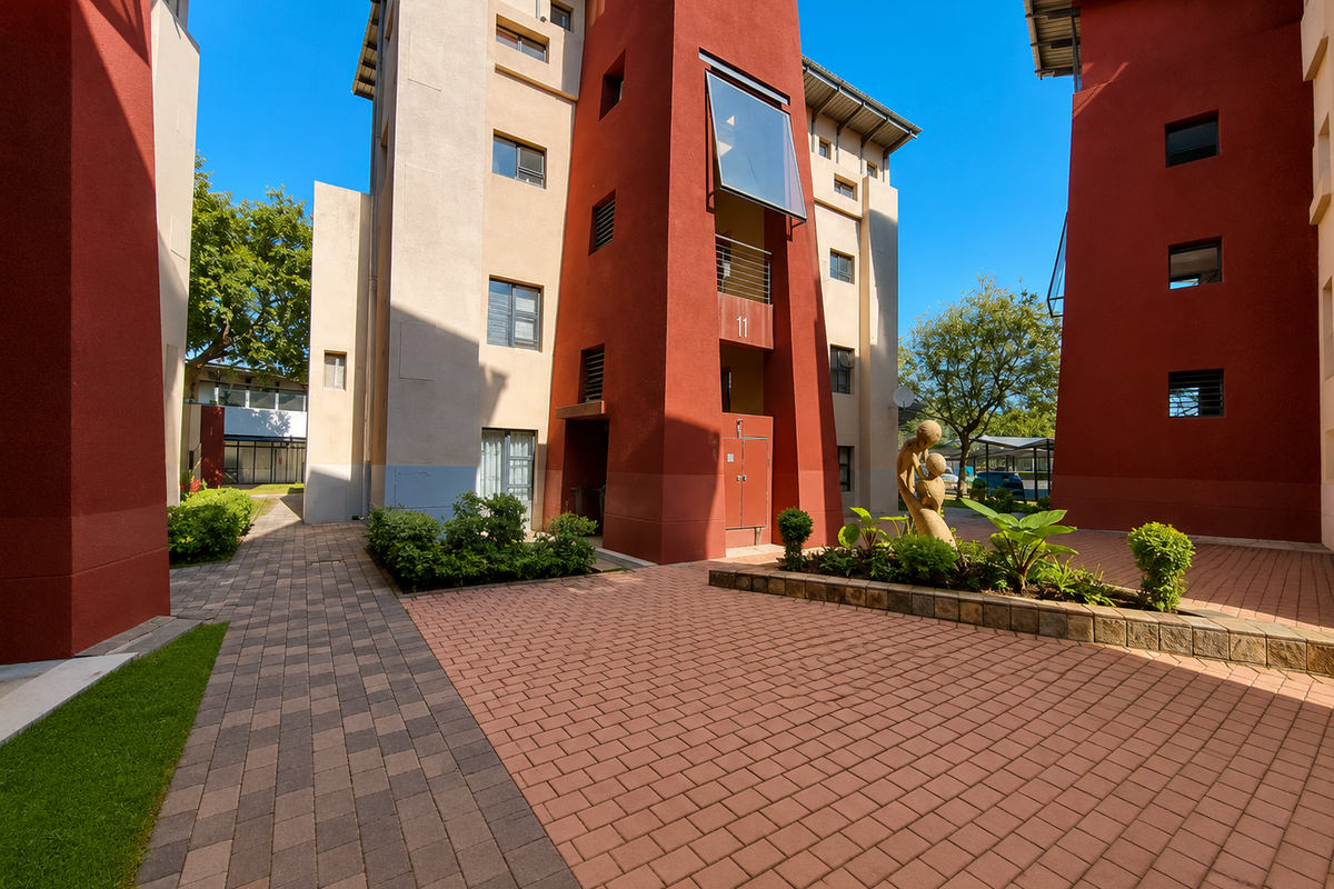 A well-maintained apartment complex courtyard featuring paved walkways, landscaped garden beds, and modern architectural facades with red and beige stucco finishes.