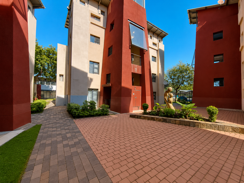 A well-maintained apartment complex courtyard featuring paved walkways, landscaped garden beds, and modern architectural facades with red and beige stucco finishes.