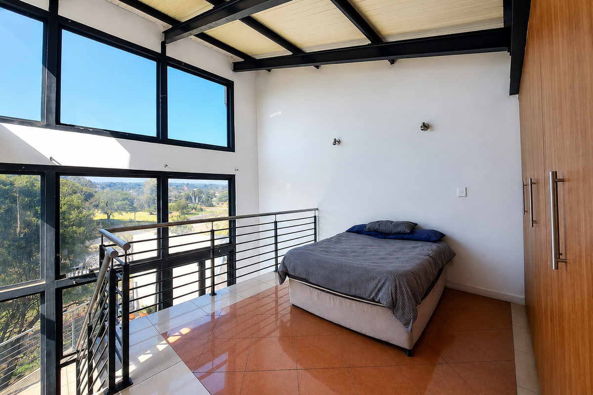 A bright mezzanine bedroom featuring high ceilings with exposed steel beams, large windows with scenic views, tiled flooring, and extensive built-in wooden cabinetry.