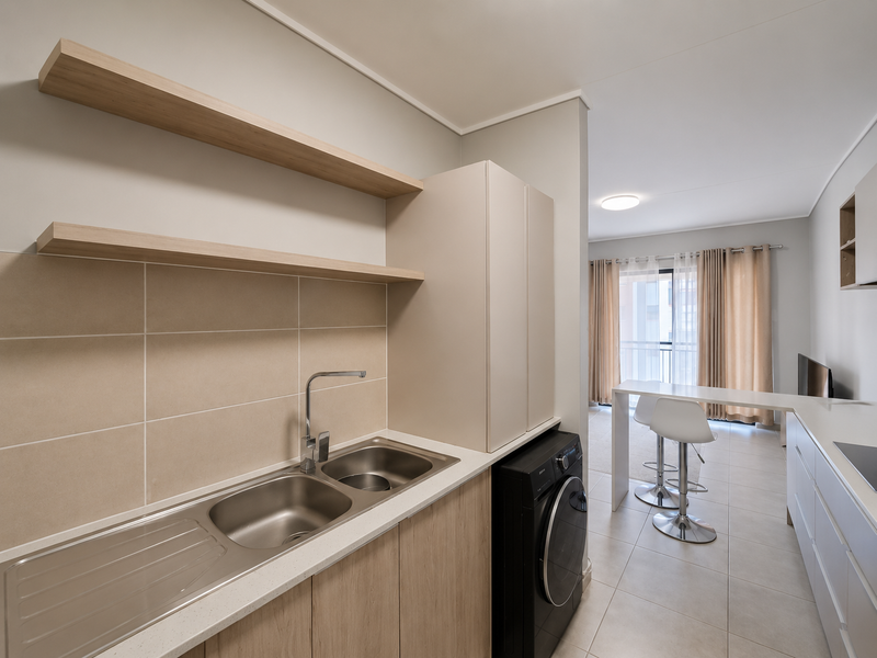 Modern open-plan kitchen area featuring sleek cabinetry, stainless steel sink, tiled splashback, and floating shelves, seamlessly flowing into the adjacent living space.