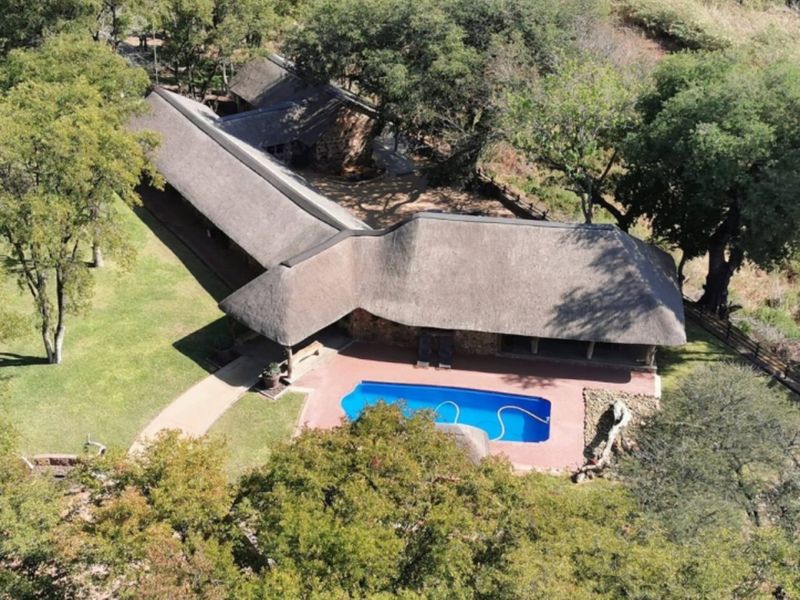 Aerial view of a farm property featuring a large thatched-roof residence, a swimming pool, and surrounding mature trees and landscaped grounds.