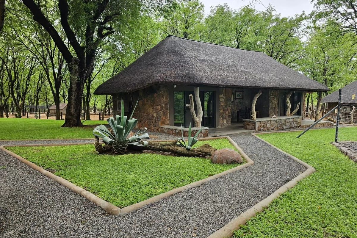 A charming stone-clad cottage with a traditional thatched roof, featuring a covered patio and surrounded by a well-maintained garden and gravel pathways.