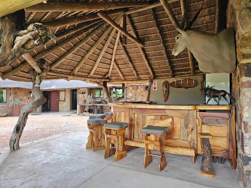 An outdoor bar area featuring a rustic thatched roof, stone wall construction, and a custom-built wooden bar counter on a concrete patio.