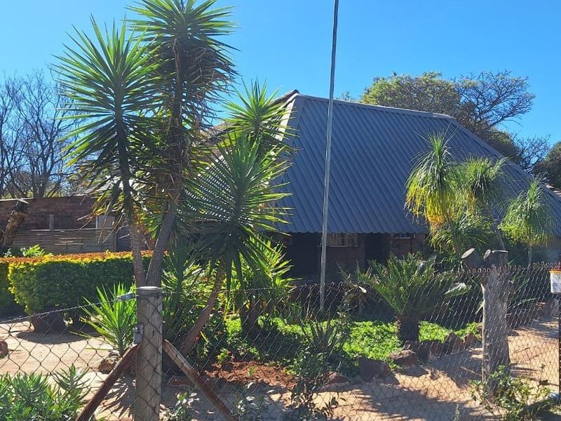 Front view of a farm-style dwelling featuring a corrugated metal roof and brick exterior, surrounded by established garden landscaping and wire fencing.