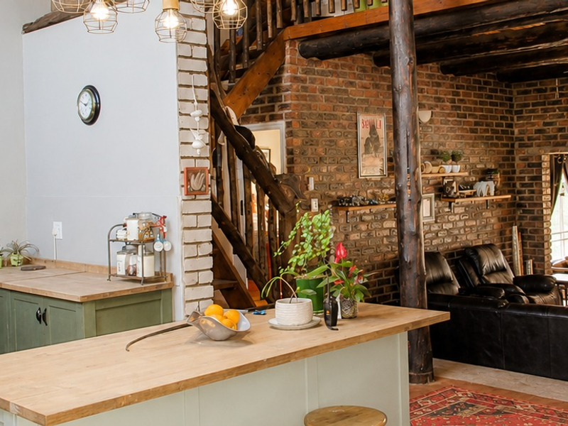 Spacious open-plan living area featuring rustic exposed brick walls, timber ceiling beams, a wooden staircase, and a functional kitchen island with wooden countertops.