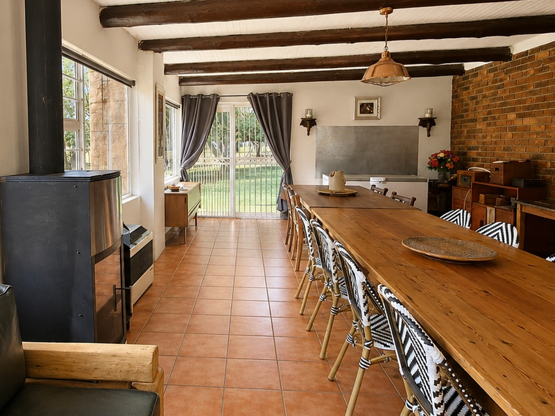 Spacious dining area featuring exposed timber ceiling beams, a rustic brick feature wall, tiled flooring, and a cozy wood-burning fireplace with garden access.