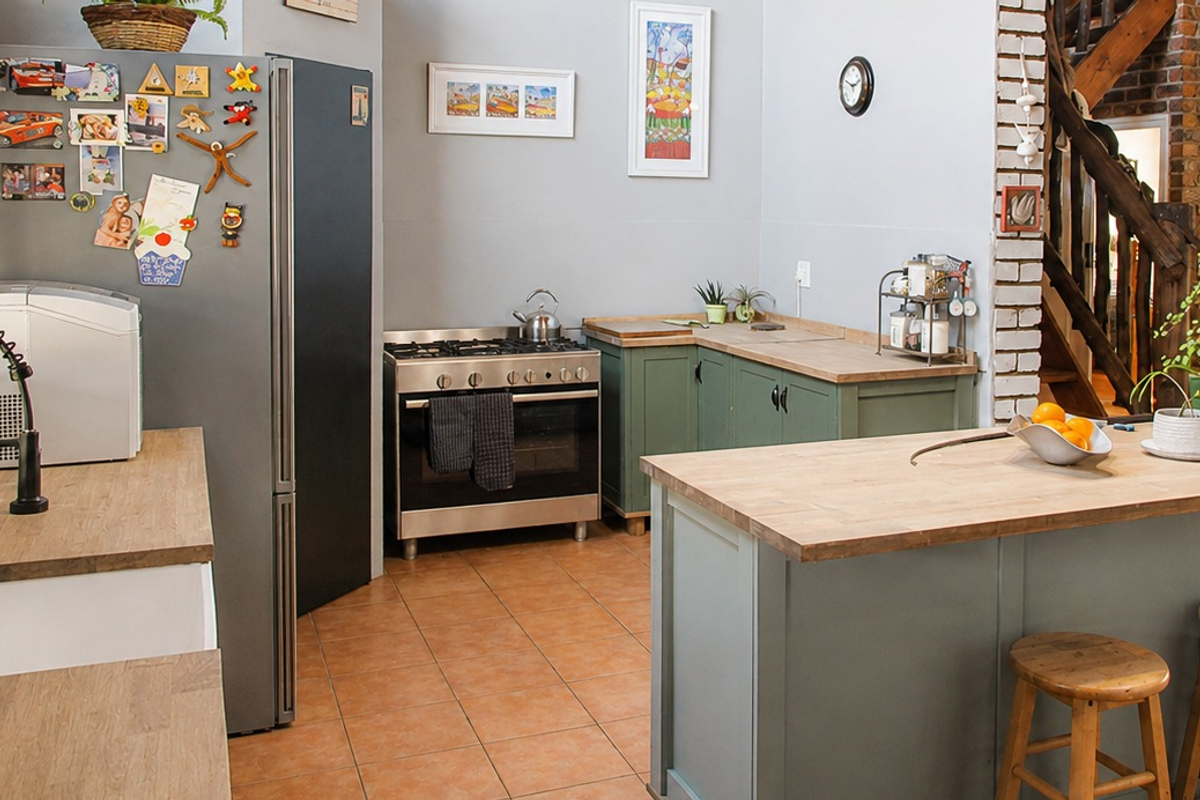 A charming farmhouse-style kitchen featuring sage green cabinetry, wooden countertops, a gas stove, and an exposed brick wall with an adjacent wooden staircase.