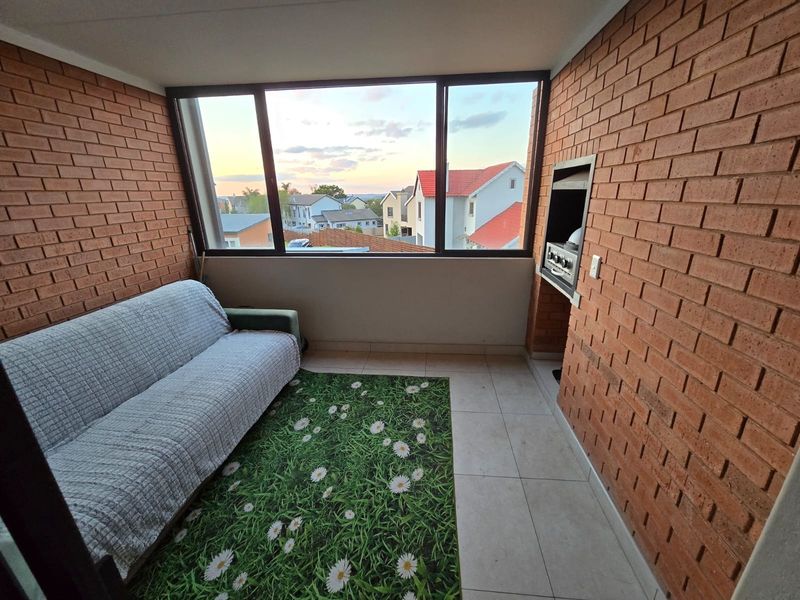 Enclosed balcony featuring exposed brick walls, tiled flooring, and a built-in braai, offering pleasant neighborhood views through large windows.