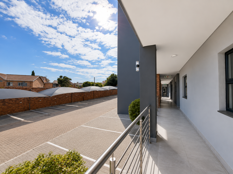 A modern covered balcony with tiled flooring and a stainless steel balustrade, overlooking a paved parking area with shade cloth carports.