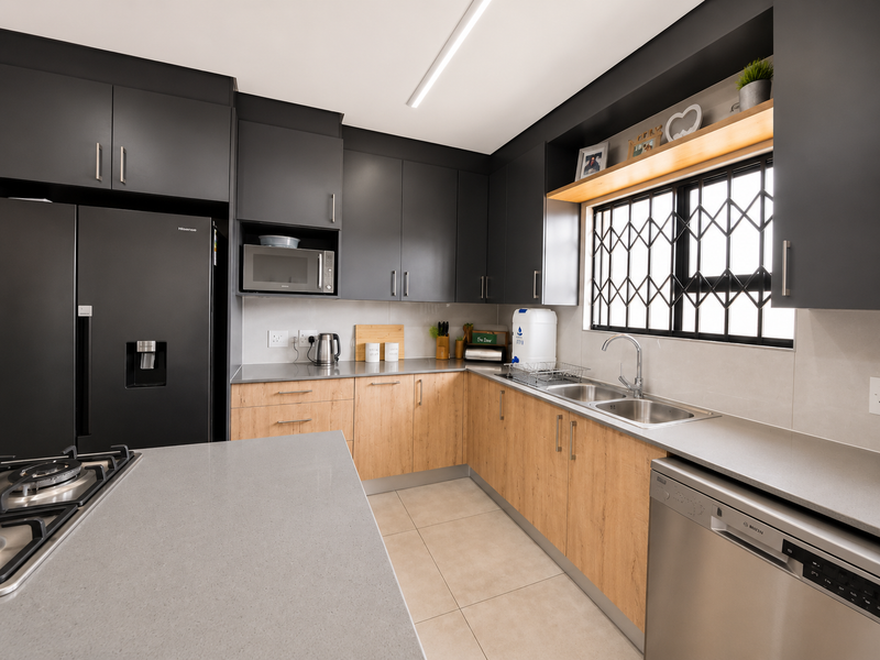 Modern kitchen featuring dark upper cabinetry, light wood-finish lower cabinets, stone countertops, integrated appliances, and a security-fitted window.