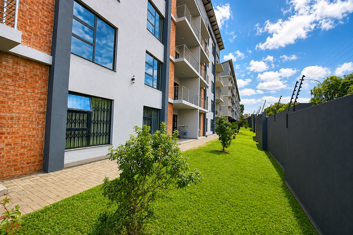 Exterior view of a modern apartment complex featuring brick and rendered facades, private balconies, a well-maintained lawn, and secure perimeter fencing.