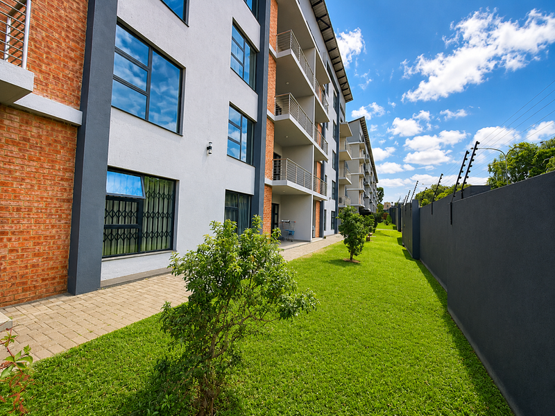 Exterior view of a modern apartment complex featuring brick and rendered facades, private balconies, a well-maintained lawn, and secure perimeter fencing.