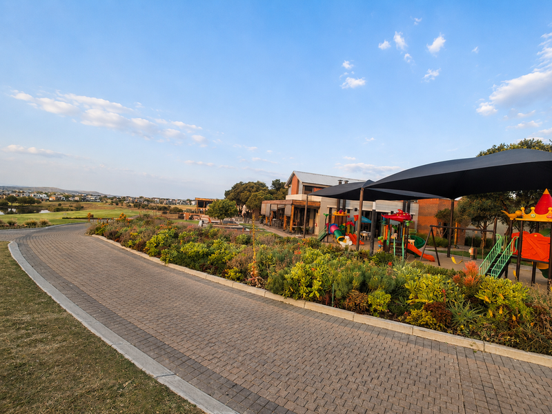 A well-maintained landscaped garden area featuring a paved walkway, shade structures, and recreational playground equipment within a residential estate setting.