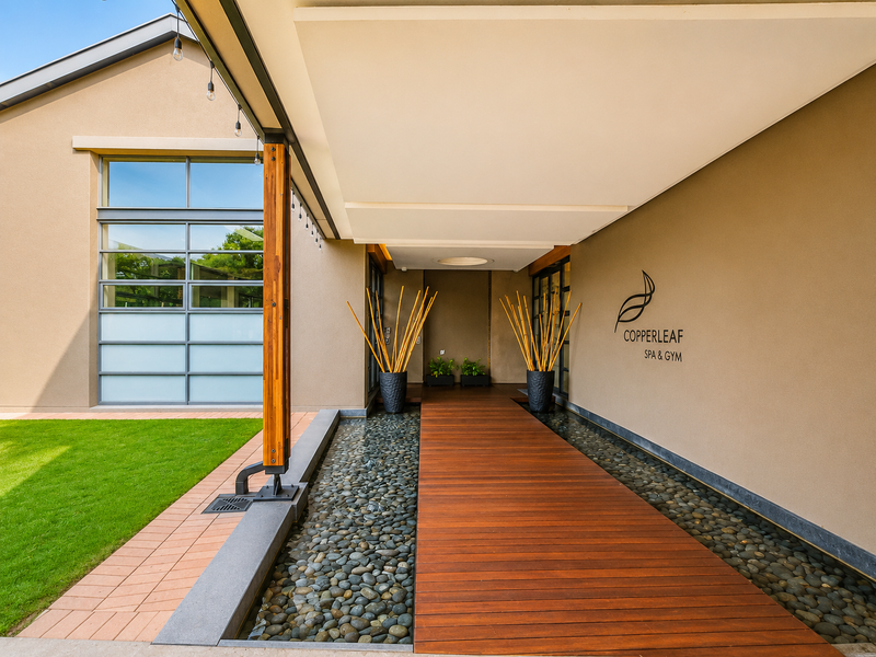 A modern entrance walkway featuring timber decking over a water feature, leading to the Copperleaf Spa and Gym facility.