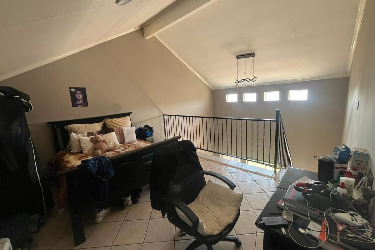 A spacious loft-style bedroom featuring vaulted ceilings with exposed beams, tiled flooring, and a metal balustrade overlooking the lower level.