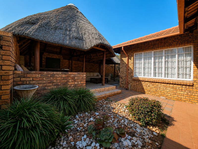 A covered outdoor patio area featuring a traditional thatched roof, built-in braai, and brick construction, adjacent to a landscaped garden with paved pathways.