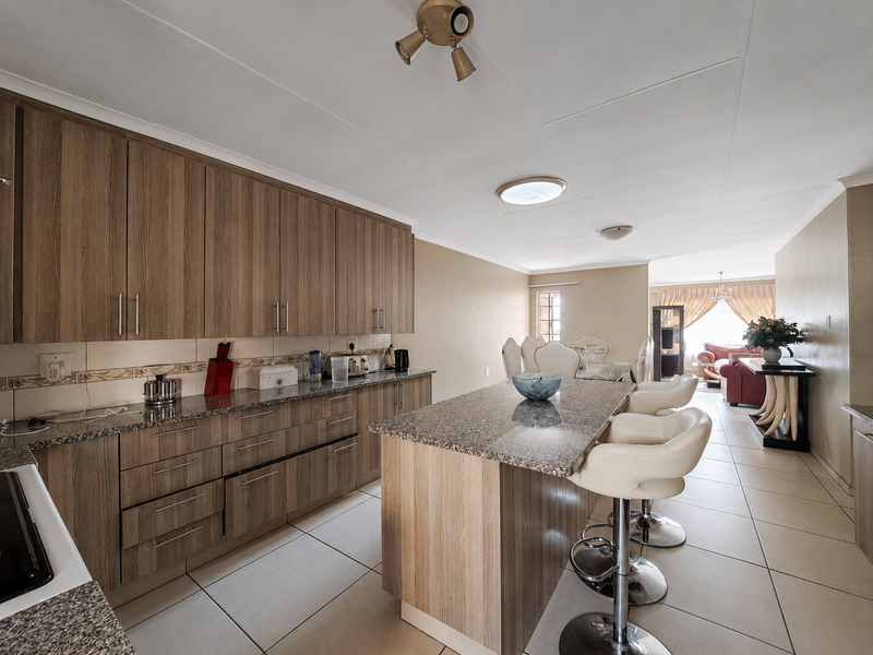 Open-plan kitchen and living area featuring wood-look cabinetry, granite countertops, a central island with breakfast bar seating, and neutral tiled flooring throughout.