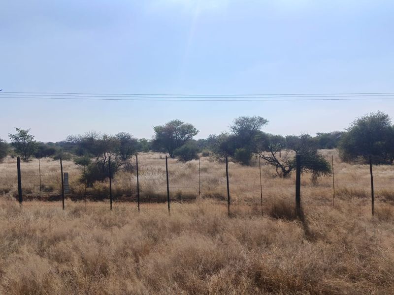 A view of a vacant residential plot featuring dry grass, native trees, and perimeter wire fencing with overhead power lines.