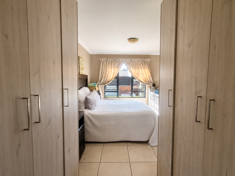 A well-lit bedroom featuring light wood-finish built-in cupboards, neutral tiled flooring, and a large window with natural light.