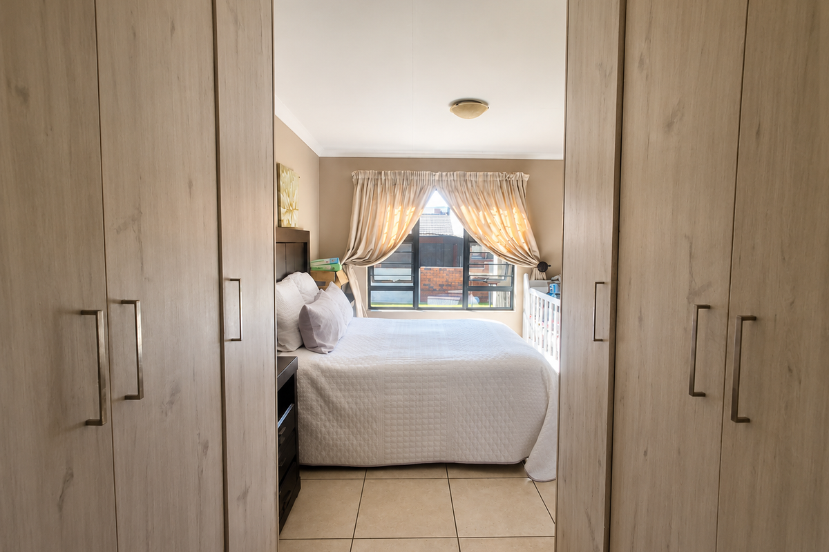 A well-lit bedroom featuring light wood-finish built-in cupboards, neutral tiled flooring, and a large window with natural light.