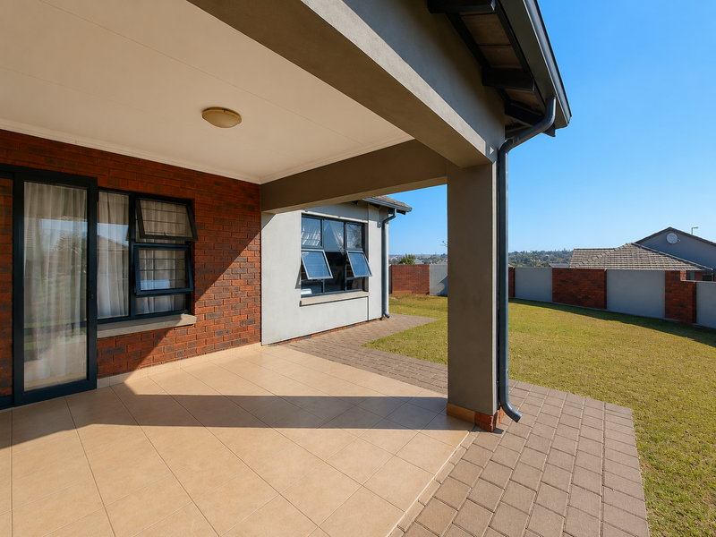 A clean, covered patio area with tiled flooring, overlooking a well-maintained garden and paved walkway, featuring modern brick and plaster exterior finishes.
