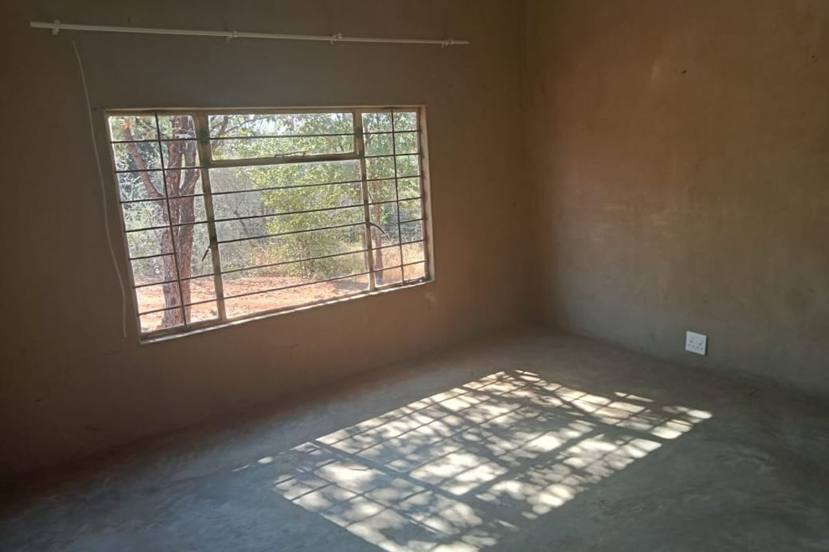 An unfinished bedroom space featuring raw plaster walls, a concrete floor, and a steel-framed window with security bars.
