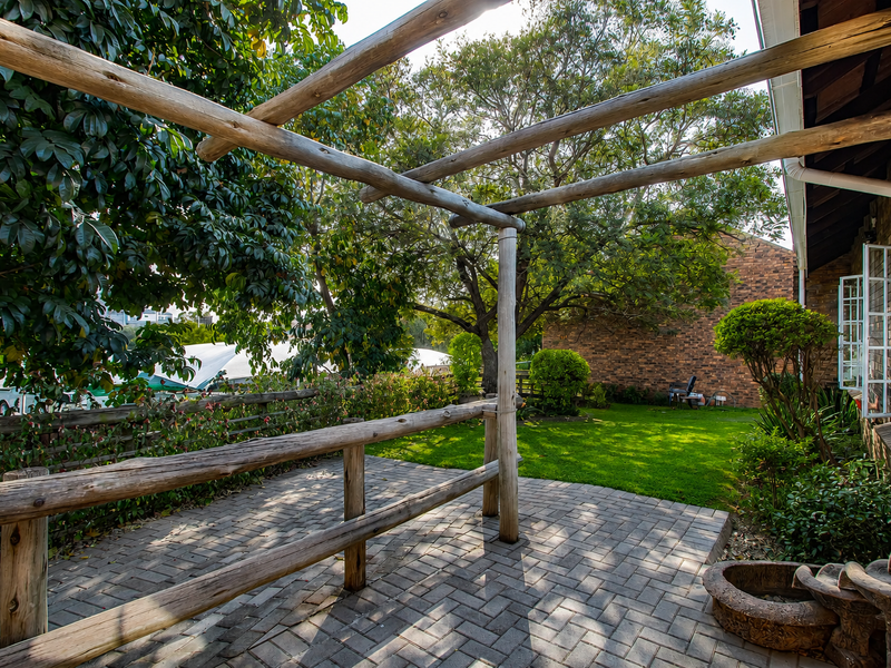 A paved outdoor patio area featuring a rustic timber pergola, post-and-rail fencing, and a stone water feature, set against a landscaped garden.