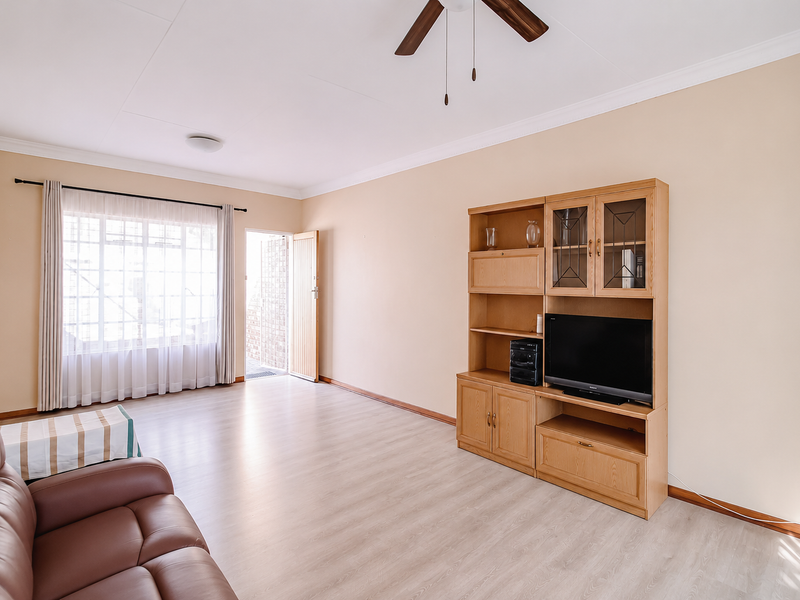 Spacious living room featuring light laminate flooring, coved cornicing, a ceiling fan, and natural light through a large window with security bars.