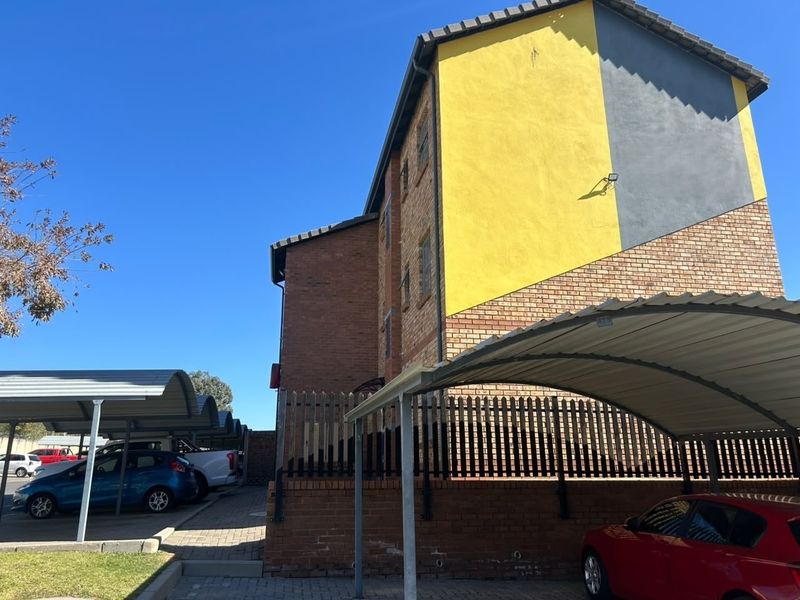Exterior view of the apartment building featuring brick facade, covered parking carports, and secure perimeter fencing.