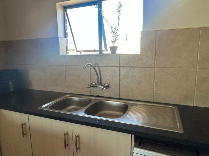 A functional kitchen area featuring a double bowl stainless steel sink, wall-mounted mixer tap, dark laminate countertops, and neutral ceramic wall tiling.