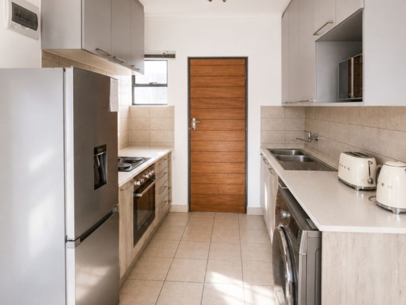 A modern galley-style kitchen featuring light wood-look cabinetry, stone countertops, integrated appliances, and neutral tiled flooring with a clean, functional layout.