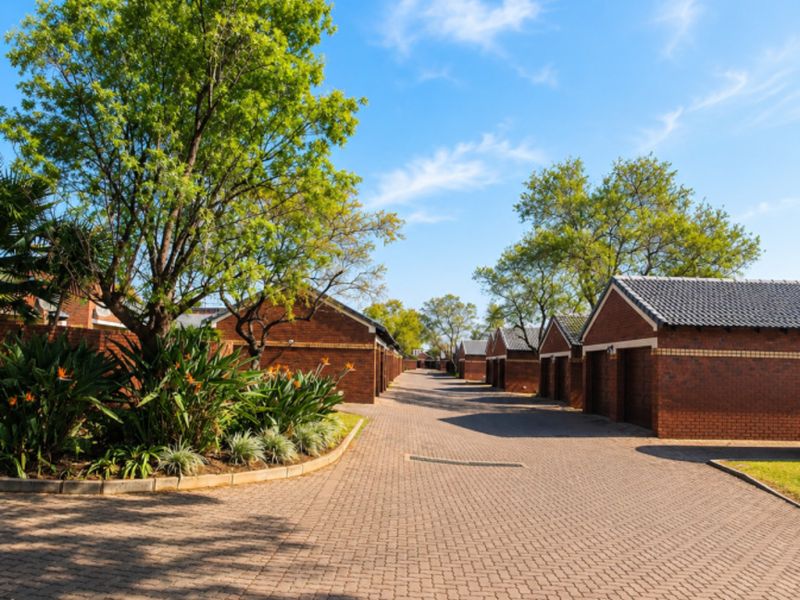 A view of the townhouse complex driveway featuring brick-paved paths, established trees, and a row of garages with tiled roofs.