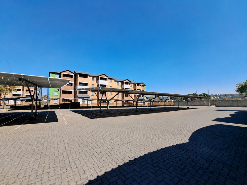 A wide-angle view of the apartment complex exterior, featuring a large paved parking area with multiple covered parking bays.