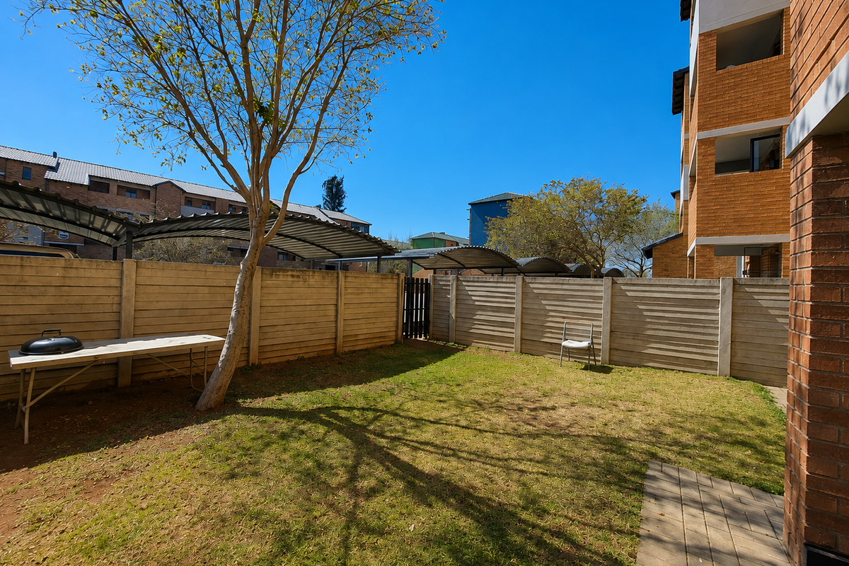 A private rear garden area featuring a lawn, paved walkway, and precast concrete boundary walls, adjacent to the apartment building and carport structures.