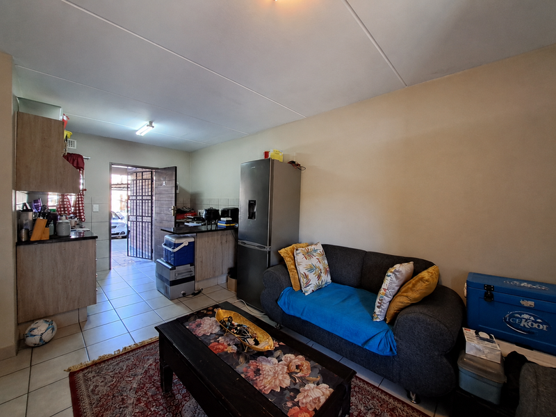 Open-plan living area featuring tiled flooring and a compact kitchen with built-in cabinetry, providing direct access to the exterior through a security gate.