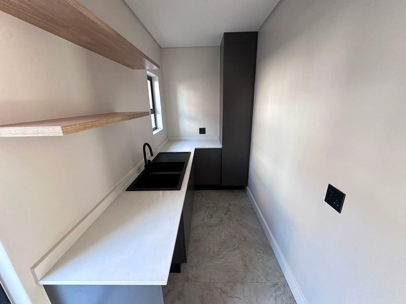 A modern, minimalist scullery featuring sleek dark cabinetry, stone countertops, a double sink with black fixtures, and floating timber shelving.
