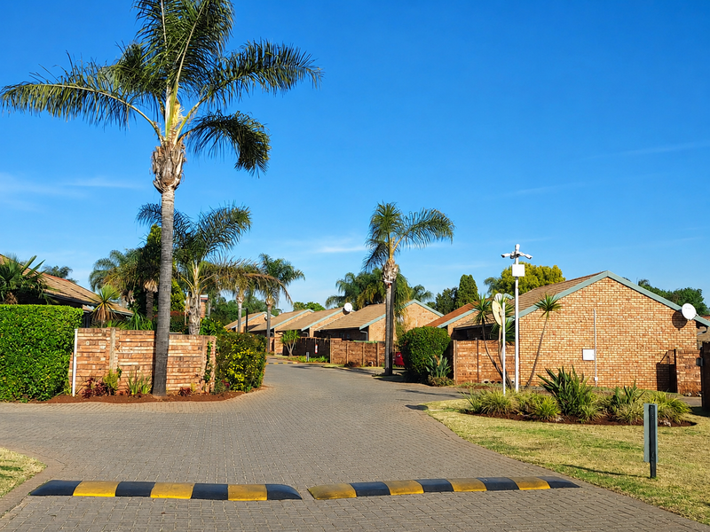 A view of the townhouse complex driveway featuring paved surfaces, speed bumps, brick boundary walls, and established landscaping.