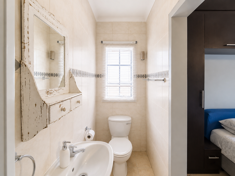 A compact bathroom featuring neutral ceramic wall and floor tiles, a pedestal basin, and a toilet, with a view into the adjacent bedroom.