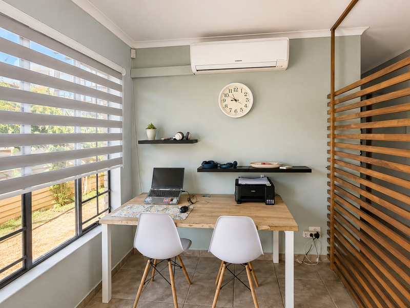 A bright home office space featuring tiled flooring, a split-system air conditioner, slatted timber room divider, and modern roller blinds.