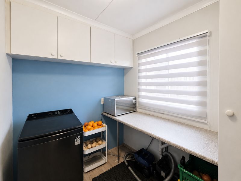 A functional laundry room featuring built-in overhead cabinetry, a spacious laminate countertop, and a window with a modern roller blind.