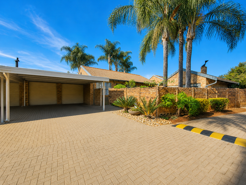 Front exterior view of the townhouse featuring a double carport, paved driveway, and established brick boundary wall with mature palm trees.