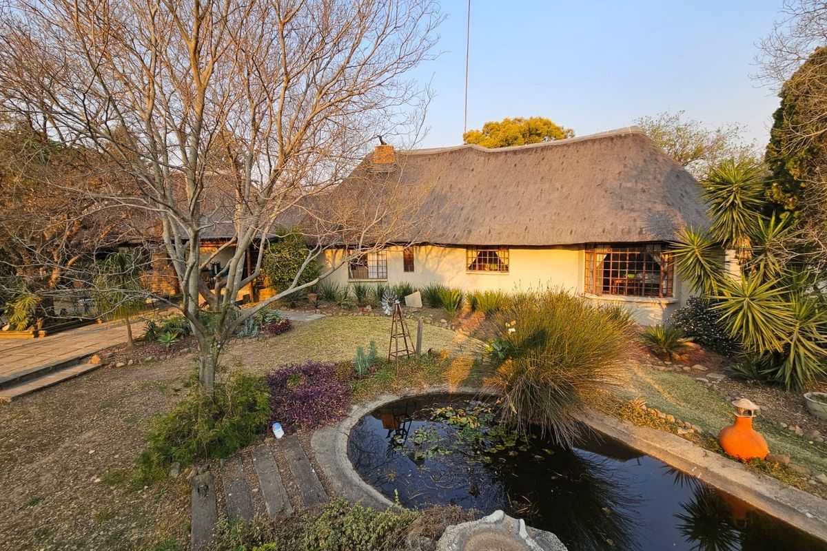 Rear view of a charming thatched-roof cottage featuring a landscaped garden with a pond and mature trees.