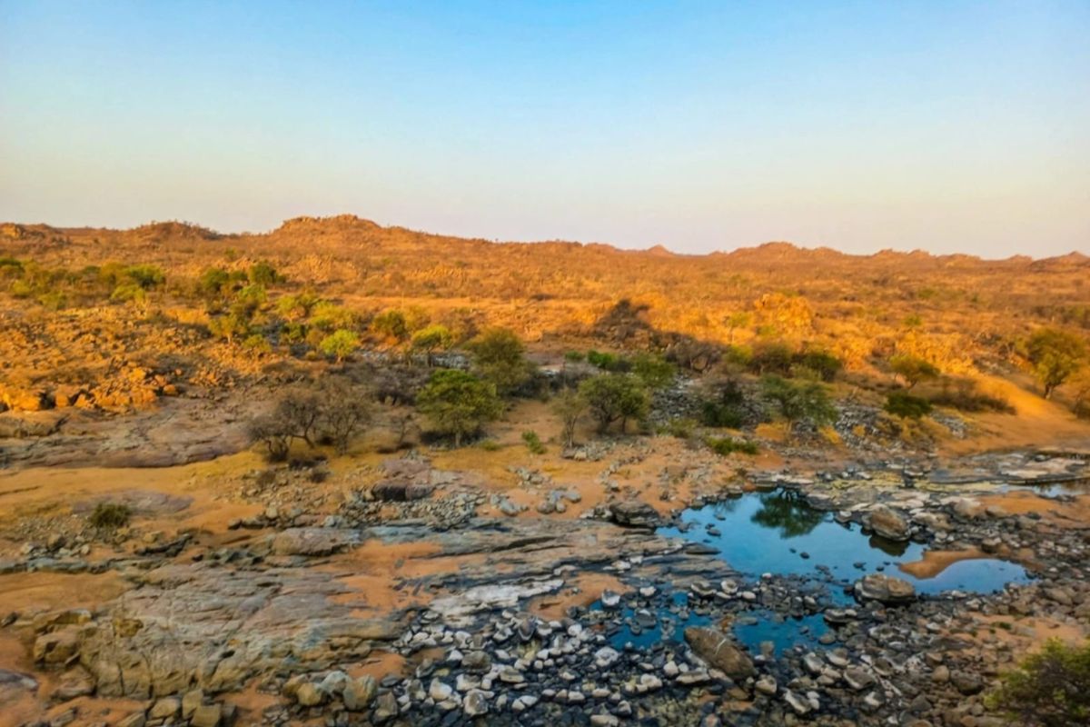 A scenic landscape view of the farm property featuring natural rock formations, indigenous trees, and a seasonal water pool under a clear sky.