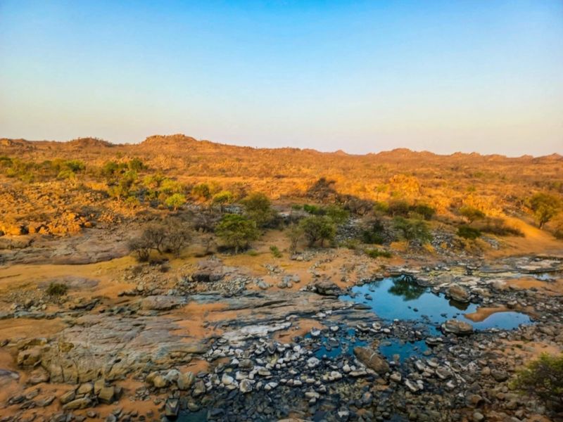 A scenic landscape view of the farm property featuring natural rock formations, indigenous trees, and a seasonal water pool under a clear sky.