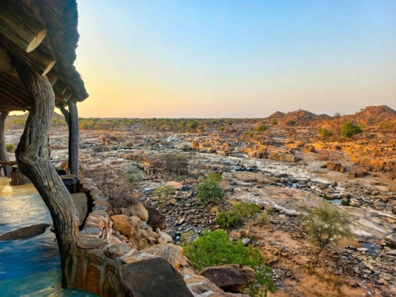 A scenic elevated view overlooking a rocky riverbed landscape from a rustic covered patio featuring natural stone masonry and timber structural elements.