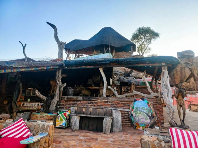 Rustic outdoor patio area featuring a thatched roof, natural wood support pillars, and stone masonry, set against a rocky landscape.