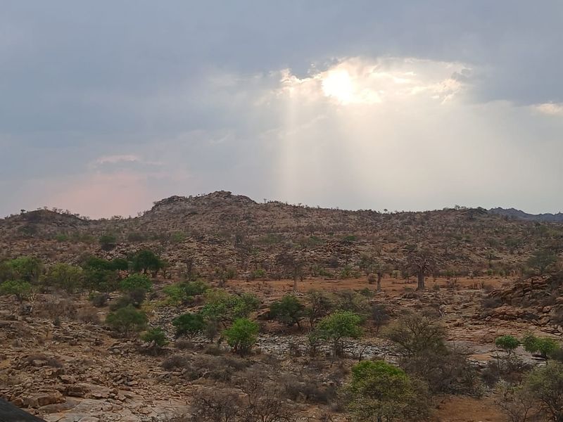A scenic landscape view of the rugged farm terrain featuring rocky outcrops, native trees, and dramatic cloud formations with sun rays.