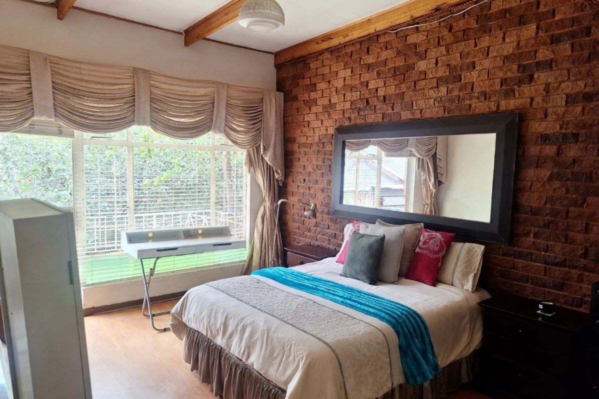 A bedroom featuring an exposed brick feature wall, timber ceiling beams, and laminate flooring, offering a rustic aesthetic with natural light.