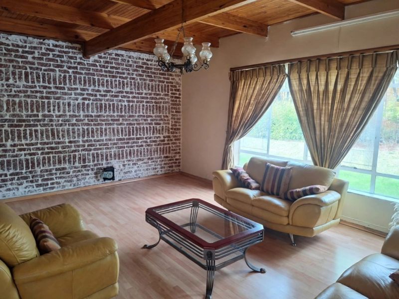 Spacious living room featuring a rustic exposed brick wall, timber ceiling beams, and large windows providing natural light.