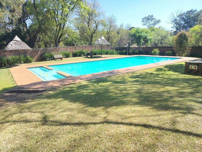 A well-maintained communal swimming pool area featuring a large pool with a shallow end, paved surround, and established garden surroundings.